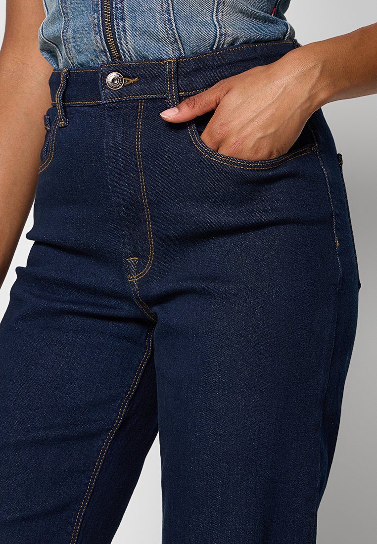 Dunkelblaue Denim-Jeans mit hoher Taille, sichtbaren Nähten und einer Vordertasche. Der Stoff wirkt robust und hat eine glatte Textur.