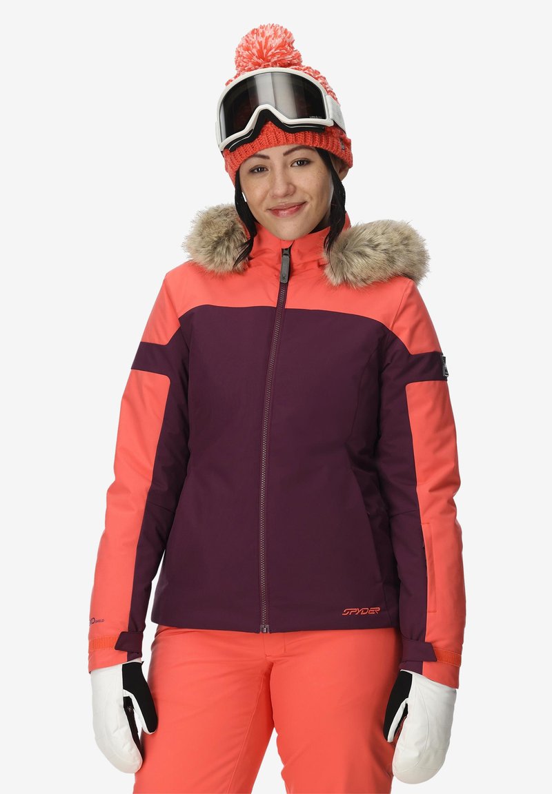 Giacca da sci viola e corallo con cappuccio in pelliccia sintetica, maniche lunghe e chiusura con zip. Abbinata a pantaloni da sci corallo coordinati e guanti bianchi.