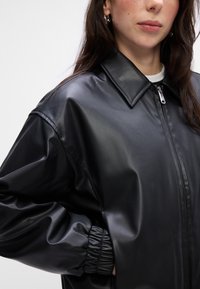 Giacca nera in ecopelle con colletto, zip frontale e polsini elasticizzati. Testura liscia con un design a vestibilità rilassata.