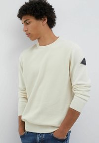 Maglione color crema con un design testurizzato e polsini a costine, caratterizzato da una patch nera a forma di triangolo sulla manica sinistra.