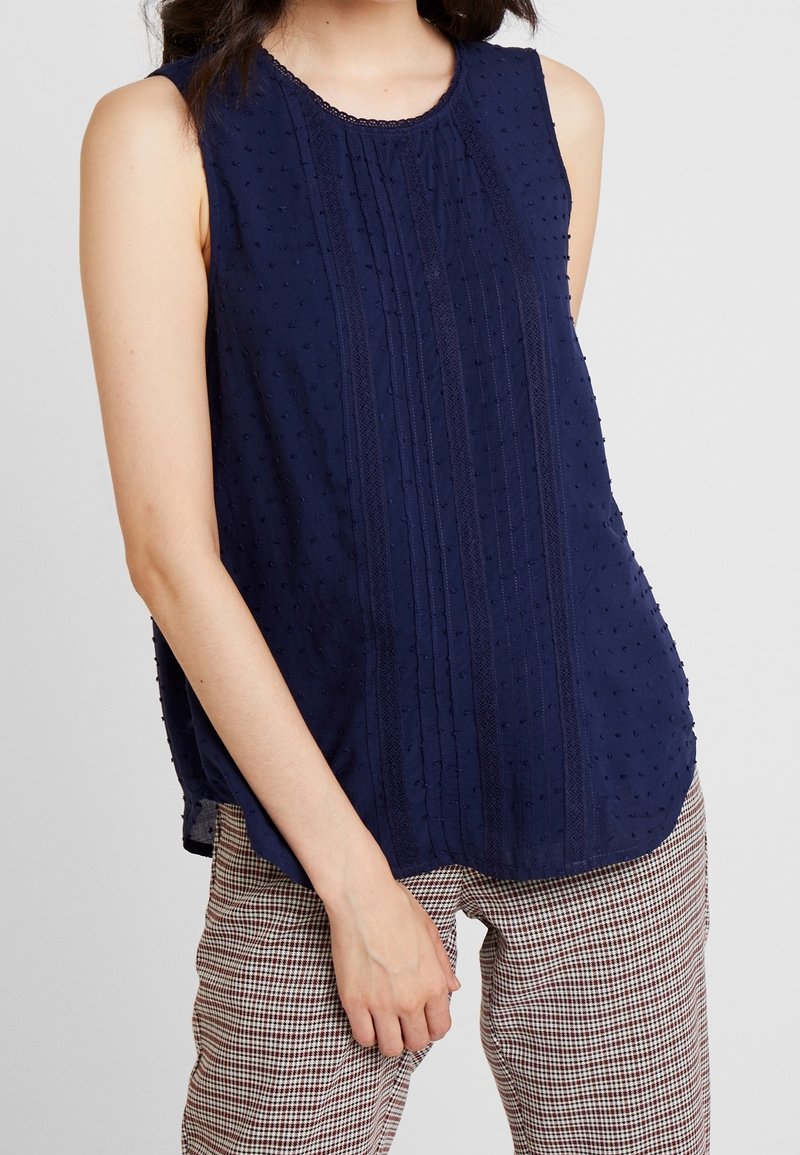 Femme portant un chemisier sans manches bleu marine texturé avec des plis fins et des détails en dentelle, associé à un pantalon à carreaux taille haute.