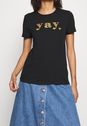 Kvinde iført en sort t-shirt med gyldent glittertekst "yay." sammen med et knapforbundet blå denimskørt, stående mod en ensfarvet baggrund.