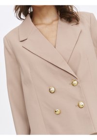 Light beige double-breasted blazer featuring a notched collar, structured shoulders, and gold button accents textured with a pattern.