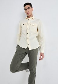 Light yellow, long-sleeve shirt with black button accents and two chest pockets, paired with olive green pants and white sneakers.