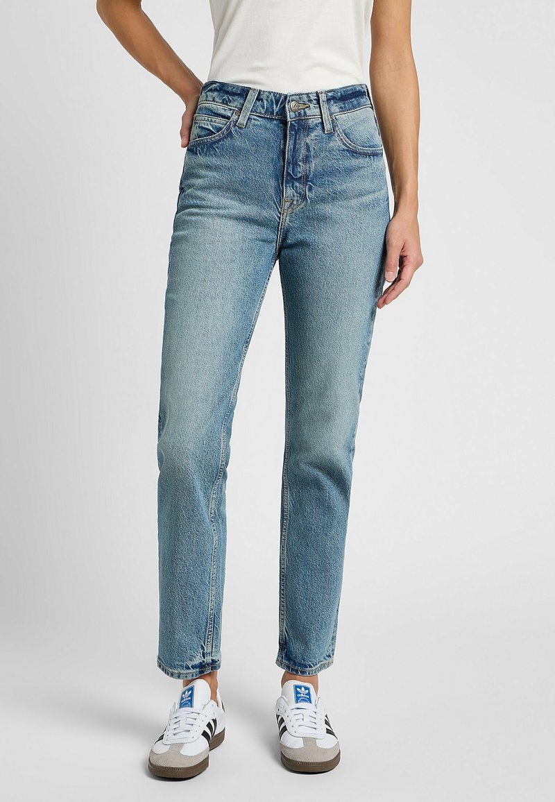 Femme portant un jean droit taille haute bleu clair et des baskets blanches avec des bandes noires et un logo bleu, debout sur un fond blanc.