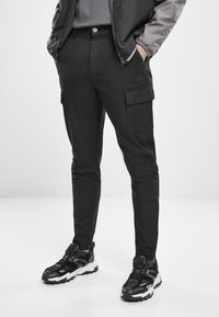 Black cargo pants with a slim fit, featuring side pockets, a button closure, and a smooth texture. Paired with black and white sporty sneakers.