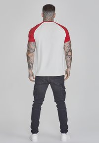 Tricou raglan roșu și alb cu mâneci scurte, asortat cu pantaloni cargo negri cu buzunare și adidași albi, văzut din spate.