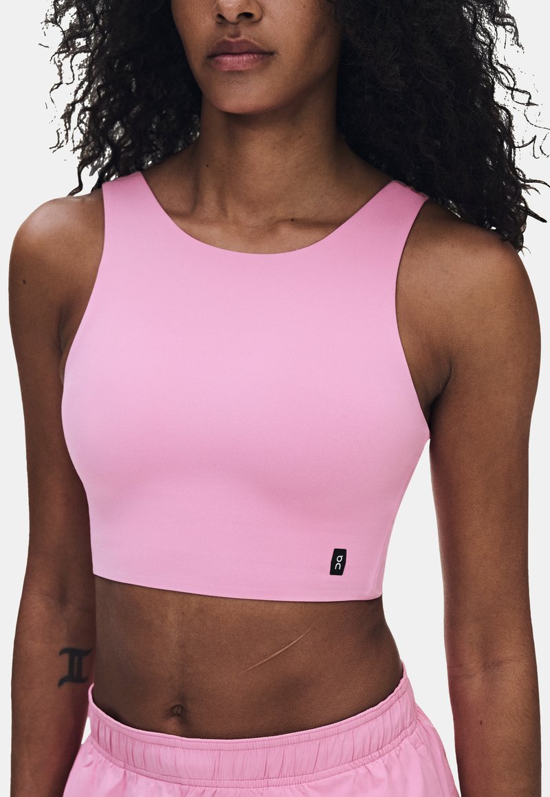 Femme portant un crop top rose clair sans manches et un short assorti, avec un petit logo sur la partie inférieure du haut.