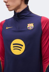 Άτομο που φοράει αθλητικό μπουφάν της FC Barcelona σε μπλε σκούρο και κόκκινο με κίτρινο λογότυπο Spotify και το σήμα της Nike στο στήθος.