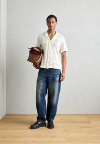 Korte mouwen, crèmekleurig gebloemd shirt, blauwe relaxte pasvorm jeans, zwarte schoenen, met een bruin leren tas met een handvat.