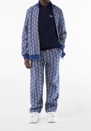 Ensemble de veste et pantalon à motifs bleus avec accents blancs. La veste présente des poignets côtelés, une fermeture éclair et un col classique. Des baskets blanches complètent le look.