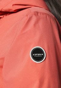 Hombro de chaqueta coral con textura suave, que presenta un parche circular en blanco y negro con el texto "ICEPEAK".