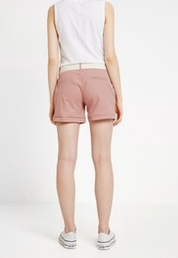 Shorts roses en tissu léger, avec un ourlet roulé et des poches arrière. Portés avec un haut sans manches blanc et des baskets blanches.