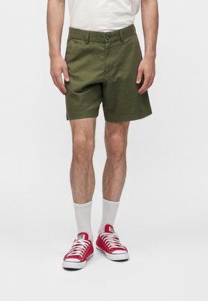 Mann trägt olivgrüne Shorts, weiße Kniestrümpfe und rote niedrige Sneaker vor einem schlichten weißen Hintergrund.
