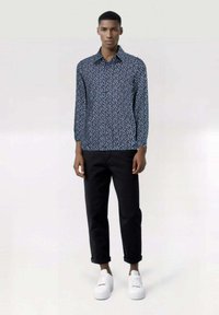 Camicia a maniche lunghe con bottoni di colore blu navy con motivo floreale bianco, abbinata a pantaloni neri e sneakers bianche su uno sfondo neutro.