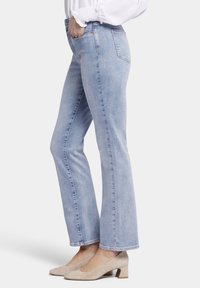 Jeans de color azul claro con corte recto, hechos de mezclilla. Se muestran con una blusa blanca y zapatos de tacón cuadrado beige, presentando un diseño clásico de bolsillo.