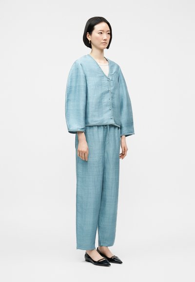 Γυναίκα όρθια, φορώντας μια ελαφριά, ανοιχτό μπλε, υφασματινή ζακέτα με κουμπιά και τζιν παντελόνι φαρδιές γραμμές, συνδυασμένα με μαύρα μυτερά παπούτσια.