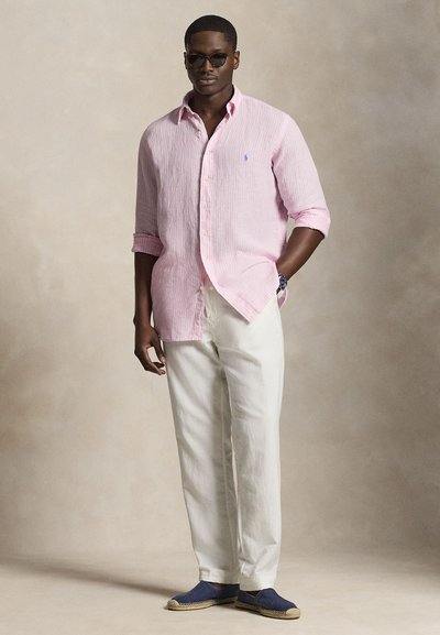 Mees, kes kannab roosat pika varrukaga särk, valgeid pükse, siniseid espadrilles'e, päikeseprille ja kella, seisab neutraalse tausta ees.