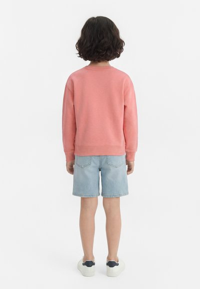 Παιδί με κοντά κατσαρά μαλλιά που φοράει ένα ροζ μακρυμάνικο φούτερ, ανοιχτόχρωμα τζιν σορτς και λευκά αθλητικά παπούτσια, στέκεται ανάποδα σε λευκό φόντο.