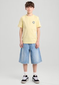 Keltainen puuvillapaita lyhyillä hihoilla ja pieni graafinen kuvio rinnassa, yhdistettynä sinisiin farkkushortseihin ja mustavalkoisiin lenkkareihin.