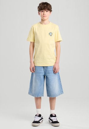 Camiseta de algodón amarilla con mangas cortas y un pequeño gráfico en el pecho, combinada con pantalones cortos de mezclilla azules y zapatillas negras y blancas.