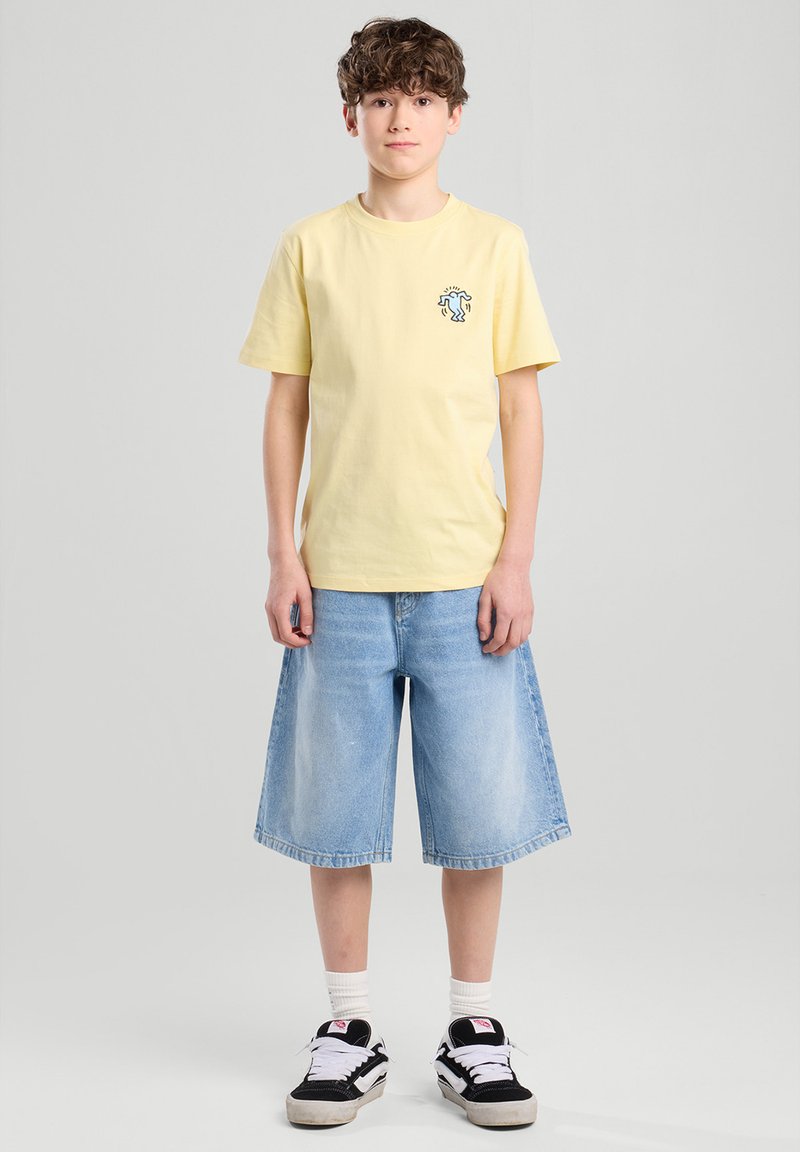 Keltainen puuvillapaita lyhyillä hihoilla ja pieni graafinen kuvio rinnassa, yhdistettynä sinisiin farkkushortseihin ja mustavalkoisiin lenkkareihin.