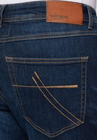 Denim jeans with a dark blue wash, featuring a leather waistband label, two back pockets with golden stitching and unique geometric designs.