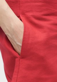 Main insérée dans la poche d'un pantalon en tissu rouge, montrant la texture et les détails de couture du vêtement.