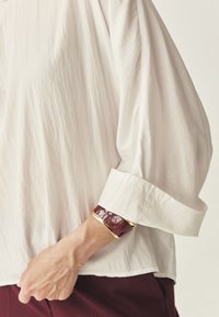 Persoon in een losse, lichtbeige blouse met opgerolde mouwen en een bordeauxrode en gouden armband om hun pols.