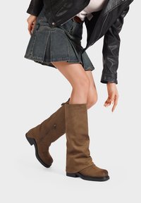 Stivali al ginocchio in suede marrone con punta rotonda e tacco basso a blocco; presentano una zip laterale e una superficie liscia. Abbinati a una gonna di jeans.