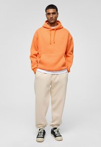 Oranje hoodie met verstelbare trekkoorden, voorzak, gecombineerd met crèmekleurige joggingbroeken en zwarte sneakers met crèmekleurige accenten.