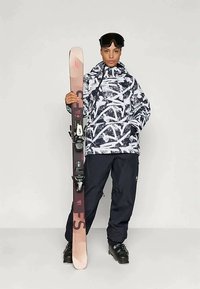 Εξοπλισμός σκι: καπιτονάτη μαύρη και λευκή τζάκετ, μαύρο παντελόνι, μαύρα μποτάκια σκι. Κρατώντας ένα ροζ και καφέ σki με μπιντάκια. Γυαλιά χιονιού στο κεφάλι.