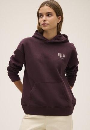 Giovane donna che indossa una felpa con cappuccio FILA Sport viola scuro con tasca frontale e pantaloni beige, in piedi con le mani dietro la schiena.
