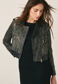 Femme portant une veste en cuir cloutée noire par-dessus une robe noire moulante côtelée, regardant vers le bas avec les cheveux légèrement soufflés sur le côté.