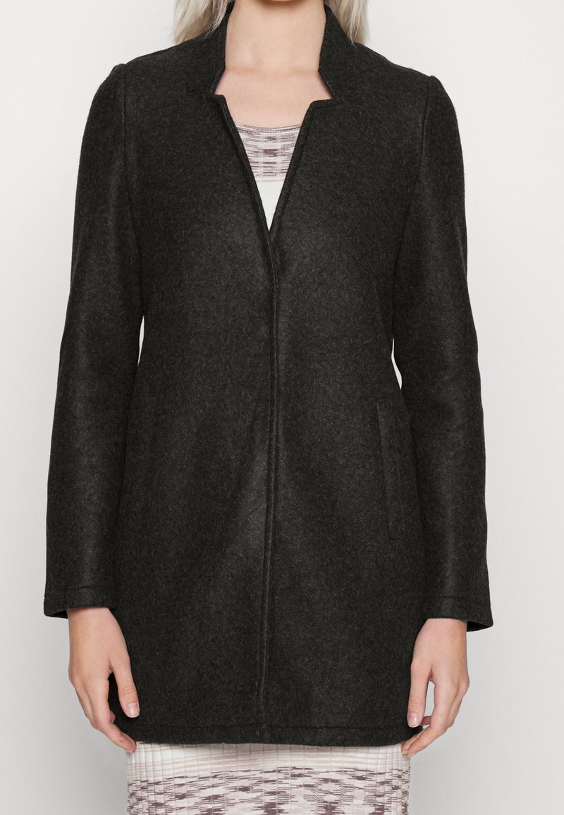 Femme portant un long manteau en laine gris charbon foncé sur une robe blanche et grise à motifs, debout avec les bras relâchés le long du corps.