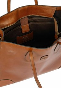 Bolso tote de cuero marrón con una textura suave, que cuenta con un interior de tela oscura, un bolsillo con cremallera y asas dobles. Etiqueta de logo visible en el interior.