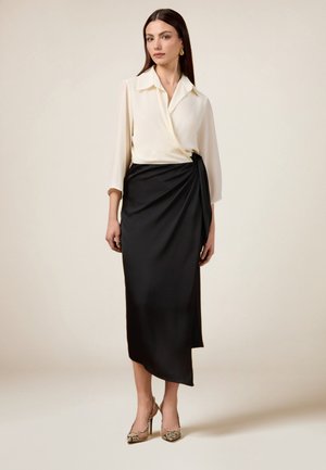 Cremfarbene Bluse mit Kragen, drei Viertelärmeln und einem Wickelrock aus schwarzem Satin mit seitlicher Bindung. Das Modell trägt hochhackige Schuhe im Tiermuster.