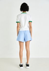 T-shirt bianca con bordi verdi, abbinata a pantaloni corti azzurri chiari. Il tessuto sembra leggero e informale. Sneakers nere e bianche completano l'outfit.