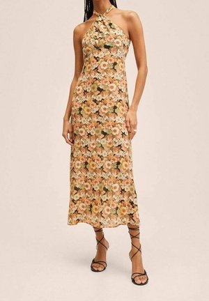 Femme portant une robe midi sans manches à col halter avec un motif floral pêche, beige et vert, et des sandales à talons hauts noires avec des lanières.