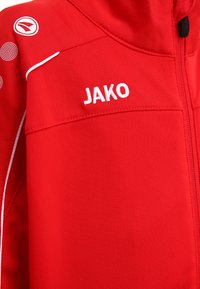 Rode sportjack met witte accenten, voorzien van een hoge kraag, een ritssluiting aan de voorkant en de tekst "JAKO" in het wit geborduurd. Stippatroon op de schouders.