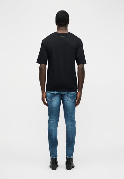 Άνδρας που στέκεται με την πλάτη στην κάμερα φορώντας μαύρο μπλουζάκι με λευκό κείμενο στο πάνω μέρος της πλάτης, μπλε τζιν και μαύρες μπότες σε απλο φόντο.