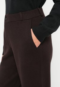 Personne portant un pantalon en tricot marron foncé avec une poche latérale et un pull noir à manches longues, la main placée à l'intérieur de la poche.