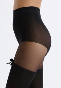 Tobilleras negras hasta el muslo con una sección transparente, parte superior sólida y un pequeño detalle de lazo en el muslo. Textura suave y diseño ajustado.