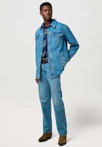 Chaqueta de denim azul claro con dos bolsillos frontales, combinada con jeans a juego y una camisa de patrón azul marino, complementada con botas de tacón marrones.