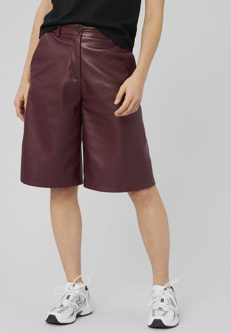 Personne portant un short en cuir bordeaux jusqu'aux genoux, un haut noir et des baskets blanches New Balance, debout contre un fond gris uni.