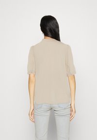 Blusa beige a maniche corte con una morbida texture, leggermente svasata sulle maniche e un colletto sobrio. Indossata sopra jeans azzurri slim-fit.