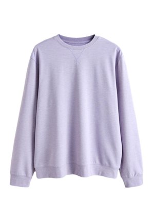 Ung mand med krøllet hår iført en lys lilla langærmet sweatshirt og blå bukser, står med den ene hånd i lommen mod en ensfarvet baggrund.