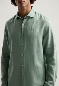 Camisa de manga larga verde claro hecha de tejido texturizado, con un frente abotonado y un pequeño logo bordado en el pecho.