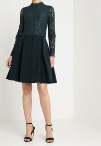 Turkos klänning med spetsliv och långa ärmar, utsvängd satinmidja, veck och svarta ankelsandaletter.