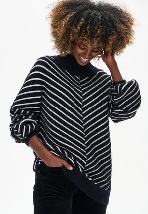 Kobieta o kręconych włosach w czarno-białym swetrze w paski i czarnych spodniach, uśmiechająca się i dotykająca włosów jedną ręką.